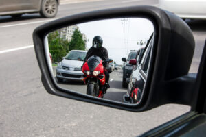 the side mirror of the car reflects a motorcyclist who is moving between cars very close to them